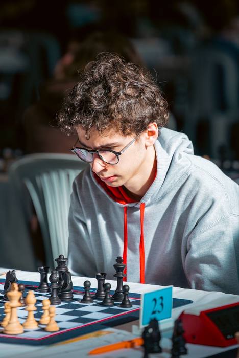 Adolescent en sweat gris penché sur son échiquier à la table 23, pendule rouge à côté